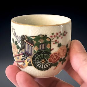Puede incluir: Una pequeña taza de cerámica blanquecina con borde dorado, que presenta una detallada ilustración de un carruaje japonés tradicional y diseños florales en tonos verdes, rojos y rosas. La taza está sostenida en una mano.