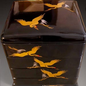 Puede incluir: Una caja hexagonal negra de varios niveles con un acabado brillante. La caja está adornada con ilustraciones de grullas doradas en vuelo, con detalles en rojo. El diseño se repite en cada nivel, creando una estética cohesiva.
