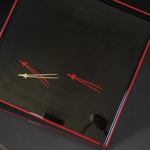 May include: A rectangular black tray with a glossy finish and a red border. The tray features two stylized red and white arrow-like designs. The tray's design is simple and elegant, suitable for serving or display.