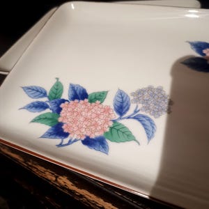 Peut inclure: Un plateau de service rectangulaire en céramique blanche orné d'un motif floral composé de grappes d'hortensias roses et bleu clair avec des feuilles vertes et bleu foncé. Le plateau présente un fin bord marron sur le pourtour.