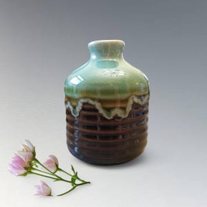 Könnte beinhalten: Eine glasierte Keramikvase in Grün- und Brauntönen, mit einem gerippten unteren Teil und einer glatten, abgerundeten Oberseite. Ein Zweig rosa Blüten liegt neben der Vase und sorgt für einen zarten Kontrast im Bild.