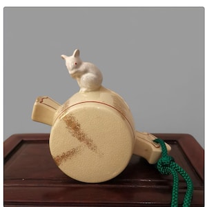 May include: A cream-colored ceramic whistle with a white rabbit figurine on top. The whistle has a circular body with brown and red accents and a green cord attached.