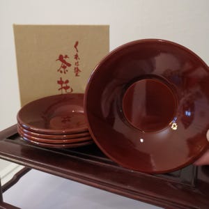Puede incluir: Un juego de seis platillos de té lacados en rojo oscuro, con un acabado brillante. Los platillos están apilados sobre una superficie de madera oscura, con una caja de cartón en el fondo con texto japonés en rojo. Cada platillo tiene una hendidura circular central.