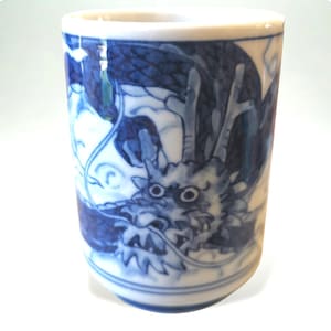 Op de afbeelding: Een cilindrische keramische beker met een wit interieur en een blauw-wit drakenontwerp. De draak is afgebeeld in een gedetailleerde, traditionele stijl, met schubben en een lang, vloeiend lichaam. De beker wordt waarschijnlijk gebruikt om thee of andere dranken te drinken.