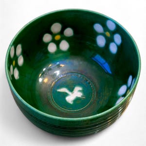 May include: A handcrafted, green ceramic bowl with white and yellow floral accents. The bowl features a white bird design in the center and a ribbed exterior. This decorative bowl is suitable for serving or display.