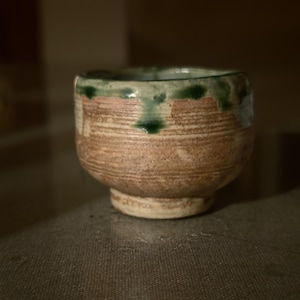 May include: A small, round ceramic cup with a textured, light brown clay body featuring horizontal ridges. The rim is decorated with a thick, uneven drip of dark green glaze that extends slightly down the sides of the vessel, which sits on a base.