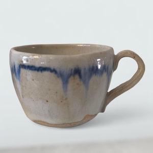 Puede incluir: Una taza de cerámica hecha a mano con un cuerpo redondeado y un asa curva. La taza es principalmente beige con un diseño de goteo azul alrededor del borde. La taza tiene un aspecto rústico y artesanal.