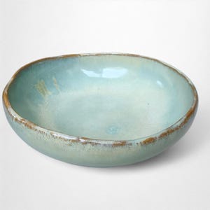 Op de afbeelding: Handgemaakte keramische kom met een lichtblauwe binnenkant en een bruine rand. De kom heeft een gladde, glanzende afwerking en een enigszins onregelmatige vorm. Geschikt voor het serveren van voedsel of als decoratief stuk. Diameter ca. 20 cm.
