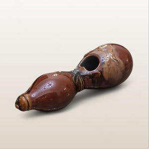 Puede incluir: Un jarrón en forma de calabaza de cerámica marrón con acabado brillante. El jarrón tiene una base bulbosa, un cuello estrecho y una parte superior redondeada con una gran abertura. Elementos decorativos son visibles en la parte superior. La calabaza es un objeto decorativo.