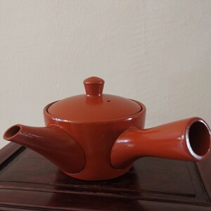 May include: A reddish-brown ceramic teapot with a lid and a side handle. The teapot has a spout on one side and a handle on the other. The lid has a small knob on top. The teapot is sitting on a dark wooden surface.