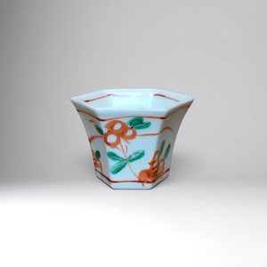 Può includere: Una tazza esagonale in ceramica bianca con motivi floreali dipinti a mano in arancione e verde. Il contenitore presenta un bordo svasato e una finitura liscia e lucida, con sottili linee arancioni che accentuano i sei angoli e il bordo superiore.