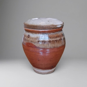 Może przedstawiać: Ceramiczny słoik z pokrywką o zaokrąglonym korpusie zwężającym się ku podstawie. Powierzchnia wykazuje gradient kolorów, przechodzący od głębokiej czerwieni terakoty na dole do nakrapianego brązowego i kremowobiałego szkliwa na górze.