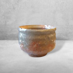 May include: A handmade ceramic bowl with a textured surface. The bowl features a color gradient, transitioning from a light gray rim to a warm orange-brown base. The bowl's exterior has a rough, organic texture.