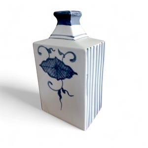 May include: A white ceramic vase with a rectangular body and a square neck. The vase features blue vertical stripes on one side and a blue floral design on the other. The top of the neck is dark blue.