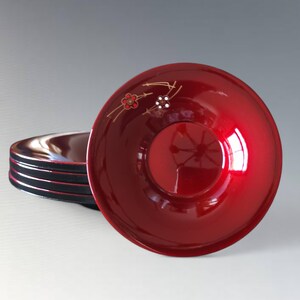 May include: A set of lacquered bowls with a deep red interior and black exterior. The top bowl features a floral design in gold and red. The bowls are stacked on a white surface.