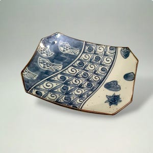May include: A rectangular ceramic plate with a blue and white design. The plate features a blue section with fish and geometric patterns, and a white section with floral motifs. The rim has a brown border.