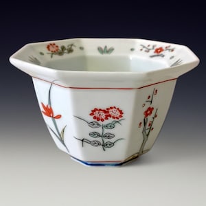 Puede incluir: Cuenco de cerámica octogonal blanco con diseños florales rojos y hojas verdes. El cuenco tiene un borde rojo y una base azul. Los diseños florales incluyen flores rojas y hojas verdes. El cuenco es probablemente para fines decorativos.