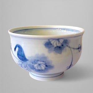 Puede incluir: Una taza de té de cerámica blanca con diseños florales azules. La taza tiene una forma redondeada con una pequeña base con pie y un borde ligeramente ensanchado. El patrón floral azul está pintado a mano.