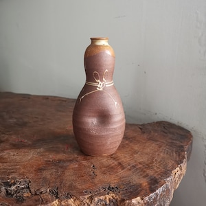 Puede incluir: Un jarrón de cerámica marrón con un diseño blanco. El jarrón tiene una forma redondeada y bulbosa y un cuello estrecho.
