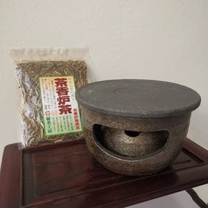 May include: A dark brown ceramic tea warmer with a lid, next to a bag of tea. The tea warmer has a round shape with a hole in the front. The bag of tea has Japanese text on it.