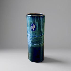 May include: A cylindrical ceramic vase featuring a glossy glaze in shades of deep navy, forest green, and teal. The surface displays a layered, drip-like pattern with a blue oval sticker near the top rim containing white Japanese text characters.