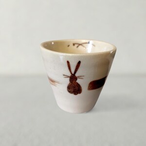 Może przedstawiać: Mały, kremowy ceramiczny kubek z brązową ilustracją królika. Kubek ma lekko rozszerzający się kształt i prosty, minimalistyczny design. Brązowe pociągnięcia pędzla zdobią zewnętrzną stronę kubka.