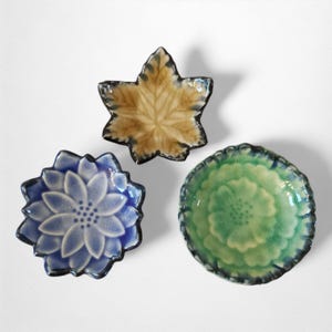 Puede incluir: Tres platos decorativos de cerámica. Uno tiene forma de hoja con tonos marrones y beige. Otro es una forma de flor azul, y el tercero es un cuenco verde con un diseño floral. Todos tienen bordes oscuros.