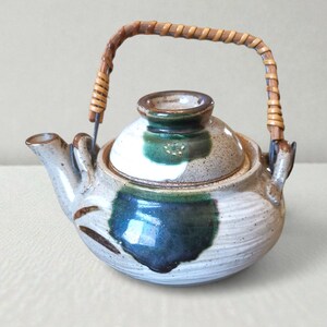 Puede incluir: Una tetera de cerámica con un asa tejida. La tetera es principalmente beige con detalles en azul oscuro y verde. El asa es de material marrón tejido. La tetera tiene tapa y boquilla.