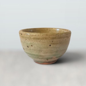 May include: A small, round ceramic bowl featuring a textured, mottled glaze in shades of beige, tan, and muted green. The surface displays a subtle crackle pattern and dark brown speckles throughout, with a smooth, rounded rim and a tapered base.