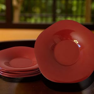 Puede incluir: Un conjunto de platos rojos y poco profundos con un círculo central elevado, junto con un solo plato. Los platos están sobre una superficie oscura, con un fondo borroso de una ventana y vegetación. Los platos tienen un acabado brillante.