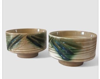 May include: Two ceramic bowls with a beige and green color scheme. The bowls have a wide, shallow shape with a rounded base and a decorative leaf pattern. The bowls are likely for serving or display.