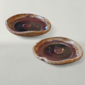 May include: Two handmade ceramic dishes. Each dish has a shallow, oval shape with a dark brown center, a red-brown ring, and a light brown outer edge. A spiral design is in the center of each dish.