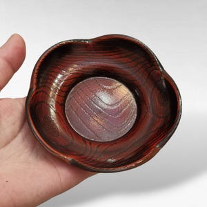 Puede incluir: Un pequeño cuenco de madera hecho a mano con un borde ondulado único. El cuenco está teñido de un color marrón rojizo oscuro, con patrones de vetas de madera visibles. El interior presenta una base circular más clara. Este cuenco decorativo es ideal para objetos pequeños.
