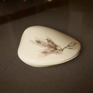May include: A smooth, off-white, pebble-shaped ceramic trinket box with a lid. The top surface features a delicate, dark brown line drawing of a pine branch with needles and a small cone, rendered in a fine, detailed illustrative style.