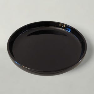 Puede incluir: Una bandeja de servicio ovalada de color negro brillante. La bandeja tiene una superficie lisa y reflectante y un borde ligeramente elevado. El color oscuro y el diseño sencillo sugieren una estética moderna, adecuada para servir alimentos o exhibir objetos decorativos.