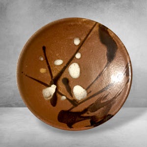 May include: A round, brown ceramic plate with a unique design. The plate features white and black abstract patterns, including splatters and linear strokes. The plate has a rustic, handmade aesthetic.