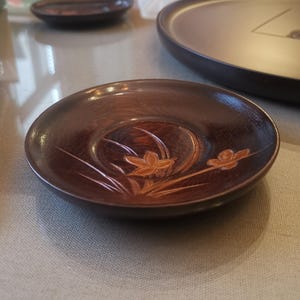 Puede incluir: Un cuenco pequeño y redondo de madera con acabado marrón oscuro. El cuenco presenta un diseño floral grabado en tonos naranja y blanco. El cuenco tiene unos 10 cm de diámetro.