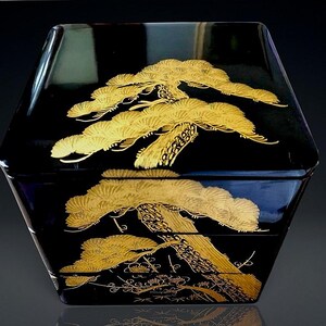May include: A square black lacquer box with a glossy finish, featuring a gold-colored pine tree design painted across the lid and the side. The intricate golden artwork depicts textured pine needles and bark, extending across the lid and base.