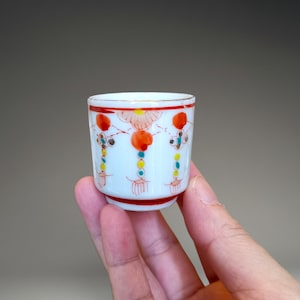 Könnte beinhalten: Kleine, weiße Keramiktasse mit handgemaltem Design. Die Tasse hat rote, orangefarbene und blaue Blumenmuster und einen goldenen Rand. Die Tasse wird in einer Hand gehalten, um ihre Größe und komplizierten Details zu zeigen. Die Tasse ist wahrscheinlich für Sake oder Tee.