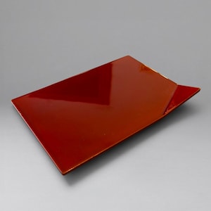 May include: A rectangular tray with a glossy, deep red finish. The tray features a flat surface with slightly angled, raised edges on one side, creating a distinct geometric profile. The material appears smooth and reflective, showing light highlights.