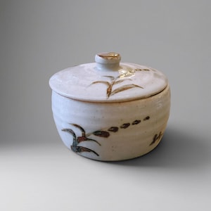 Op de afbeelding: Een ronde keramische pot met deksel, voorzien van een crèmekleurig glazuur en een gespikkelde textuur. De buitenkant is versierd met handgeschilderde bruine en groene botanische motieven van bladeren en stengels op de basis en het deksel.