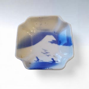 May include: A small, square, white ceramic dish with a blue mountain scene and flying birds. The dish has a scalloped edge and is painted with shades of blue and white. The design is reminiscent of traditional Japanese art.