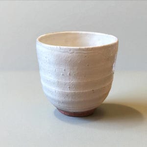 Puede incluir: Una taza de cerámica blanca hecha a mano con una superficie texturizada y crestas horizontales. La taza tiene una forma redondeada y una base marrón rojiza. El interior también es blanco, sobre un fondo neutro.