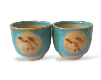 Puede incluir: Dos tazas de cerámica turquesa con base y borde beige. Cada taza presenta un círculo beige con un diseño abstracto marrón. Las tazas están una al lado de la otra sobre un fondo blanco.