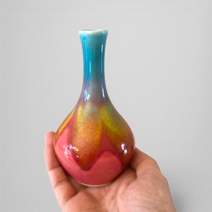 May include: A small, colorful ceramic vase with a teardrop shape. The vase has a gradient of colors, starting with a teal blue at the top, transitioning to gold and orange, and ending with a vibrant pink at the base. The vase is held in a hand.
