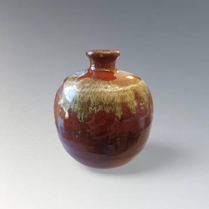 Puede incluir: Un pequeño jarrón de cerámica redondo con cuello estrecho. El jarrón es principalmente de color burdeos oscuro, con una banda de esmalte dorado y crema en el medio. El esmalte tiene un efecto goteado, agregando textura a la pieza.