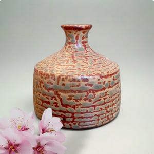 May include: A ceramic vase with a textured surface in shades of red, tan, and gray. The vase has a rounded body and a narrow neck. Pink cherry blossoms are in the foreground, adding a touch of color and contrast.