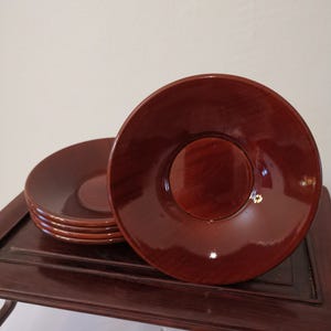 Puede incluir: Un conjunto de seis platillos de color rojo oscuro y brillante. Cada platillo es circular con un centro ligeramente elevado. Se exhiben sobre una superficie de madera oscura, adecuada para una mesa de comedor o servicio. Los platillos miden unos 15 cm de diámetro.