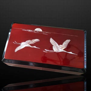 Puede incluir: Caja rectangular de laca roja con acabado brillante. La tapa presenta dos grullas blancas en vuelo, con un diseño de luna y línea de agua. La caja tiene un borde plateado y una base negra.