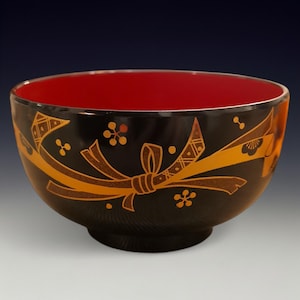 May include: A black bowl with a red interior. The exterior is decorated with gold-colored ribbon and floral designs. The bowl has a rounded shape and sits on a small, circular base. The bowl is likely made of ceramic or a similar material.